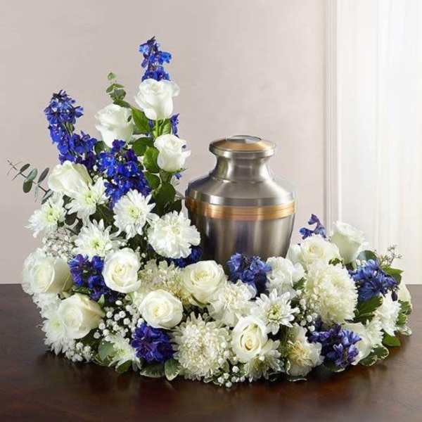 Cremation urn ringed by white roses, white mums, and blue flowers in a low sympathy arrangement.