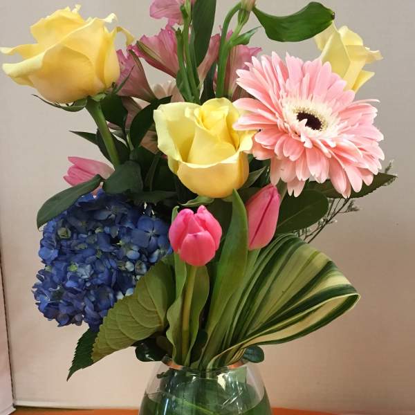 Mixed bouquet in a clear glass vase with yellow roses, pink gerbera, blue hydrangea, and tulips