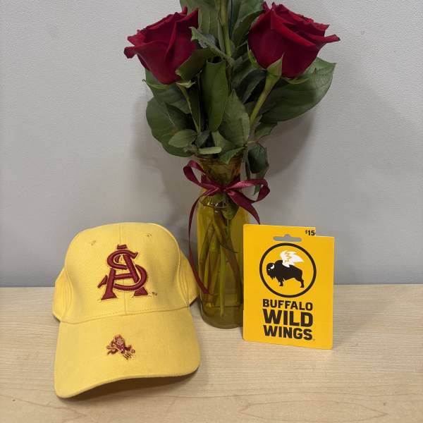 Three red roses in a glass vase with a ribbon, beside a yellow cap and gift card.