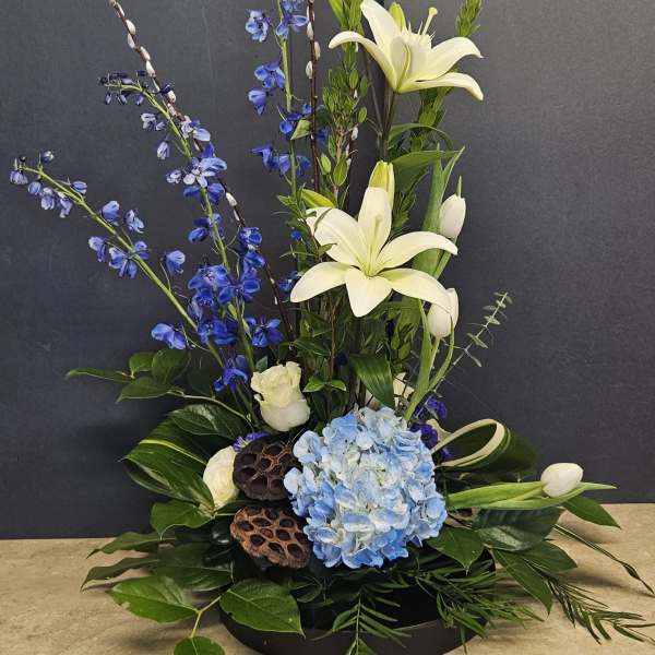 Tall floral arrangement with white lilies, blue flowers, and hydrangea in a low black base