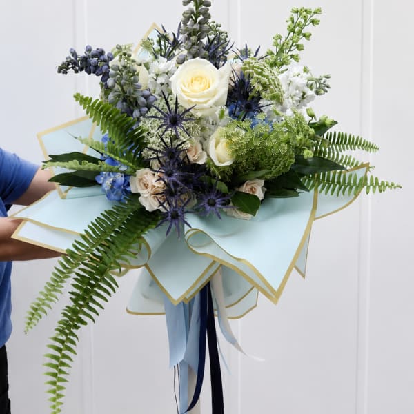 Handheld bouquet of white roses, blue flowers, and fern fronds wrapped in pale blue paper