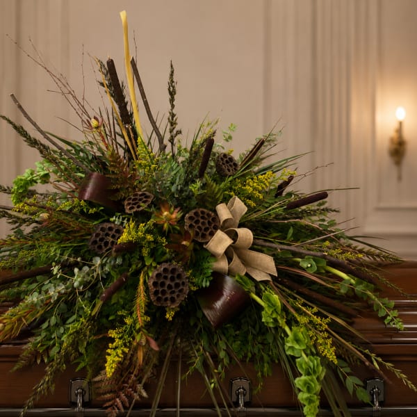 Large funeral spray with greenery, lotus pods, and beige ribbon on a casket