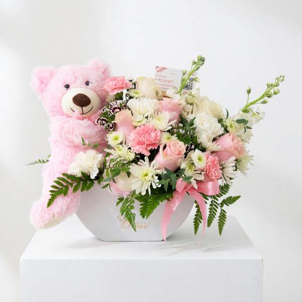 Pink teddy bear beside a pastel flower arrangement in a white box