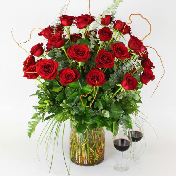Red roses arranged in a glass vase with greenery and two wine glasses