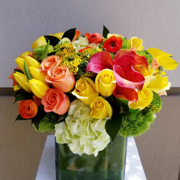 Bright bouquet of yellow roses, pink calla lilies, and orange flowers in a square vase