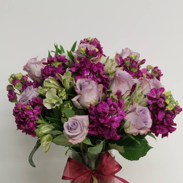 Round arrangement of lavender roses and bright purple flowers in a clear vase with a pink bow