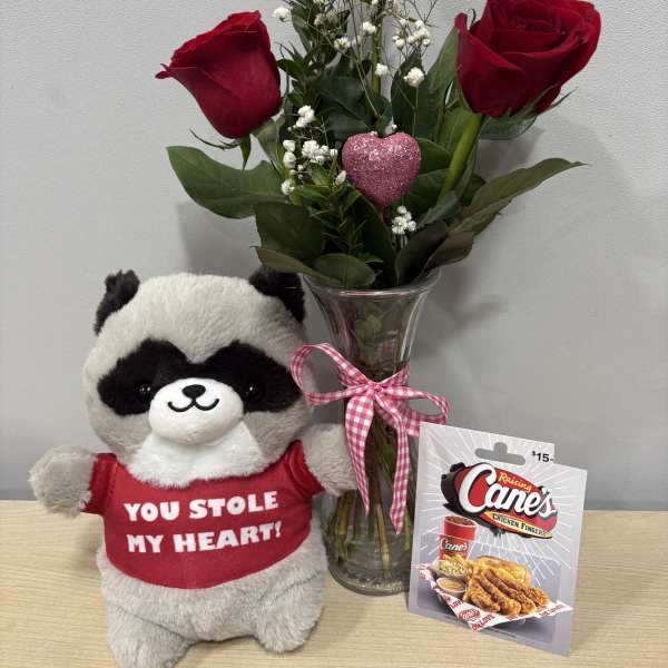 Three red roses in a glass vase beside a plush raccoon and gift card