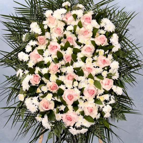 Large standing spray of pink roses and white flowers framed by palm fronds on a wooden easel.