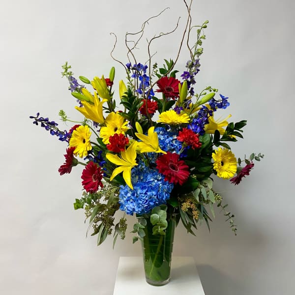 Tall mixed bouquet in a glass vase with yellow lilies, gerberas, blue hydrangea, and red blooms