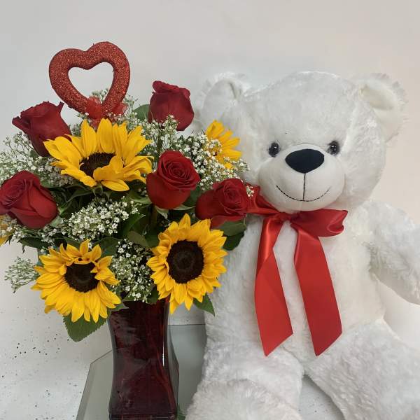Red roses and sunflowers in a vase beside a white teddy bear with a red bow