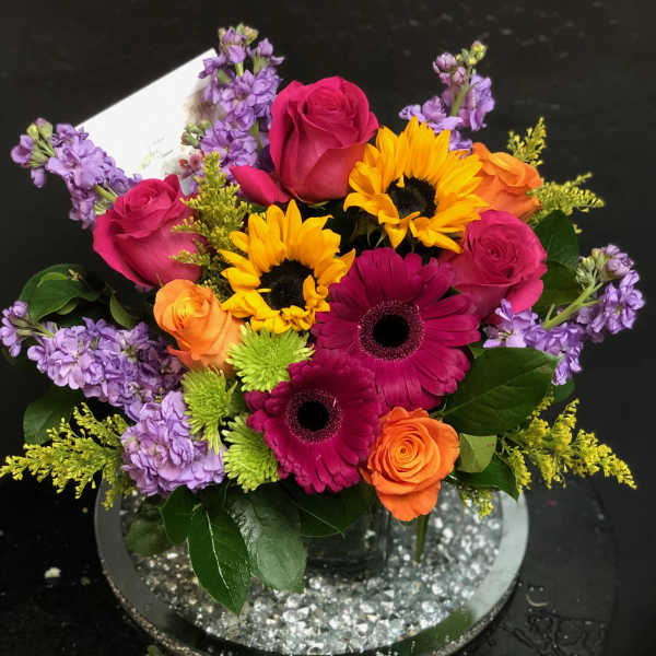 Bright mixed bouquet with roses, gerbera daisies, sunflowers, and purple blooms in a vase