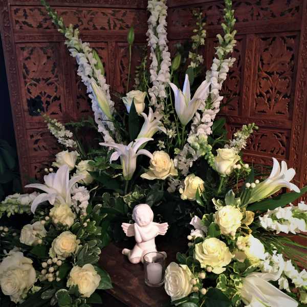 White floral funeral arrangement with lilies and roses around a small angel figurine