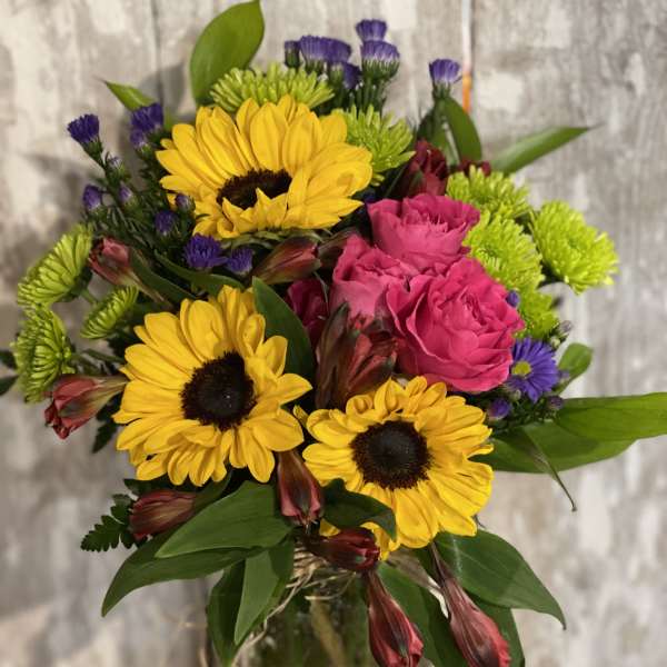 Bouquet of yellow sunflowers, pink roses, and purple filler flowers in a glass vase