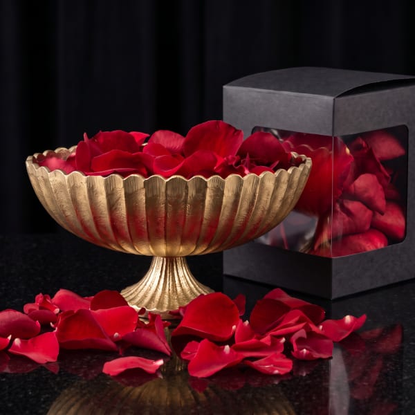 Gold pedestal bowl filled with red rose petals beside a black gift box
