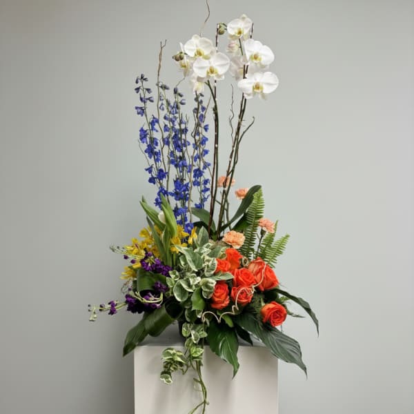 Tall floral arrangement with white orchids, blue delphinium, and orange roses