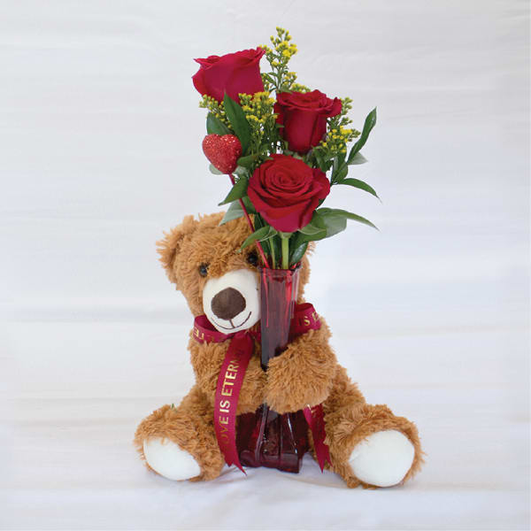 Teddy bear holding a vase of red roses with a heart pick