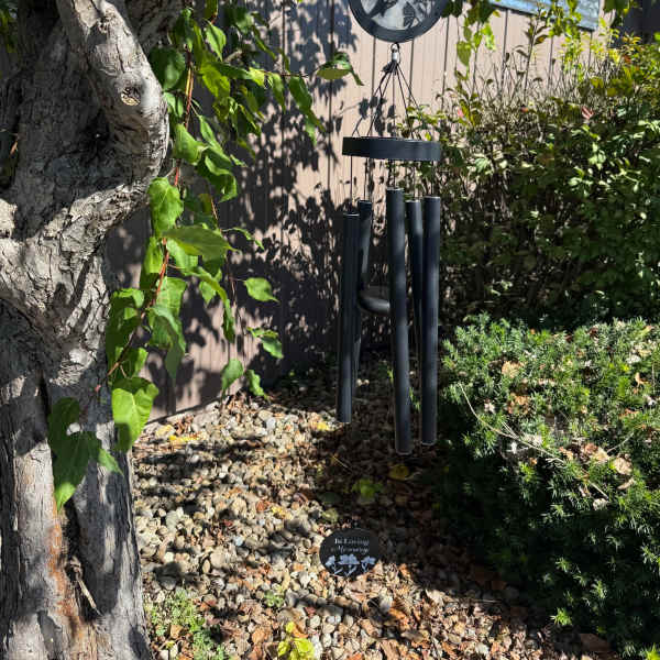 Black memorial wind chime with hummingbird design hanging from a tree over a rock garden.