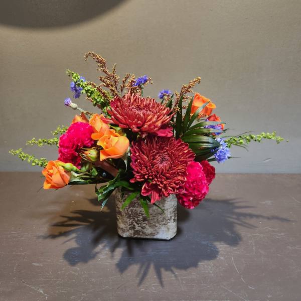 Mixed bouquet of orange roses, red chrysanthemums, and pink carnations in a square vase