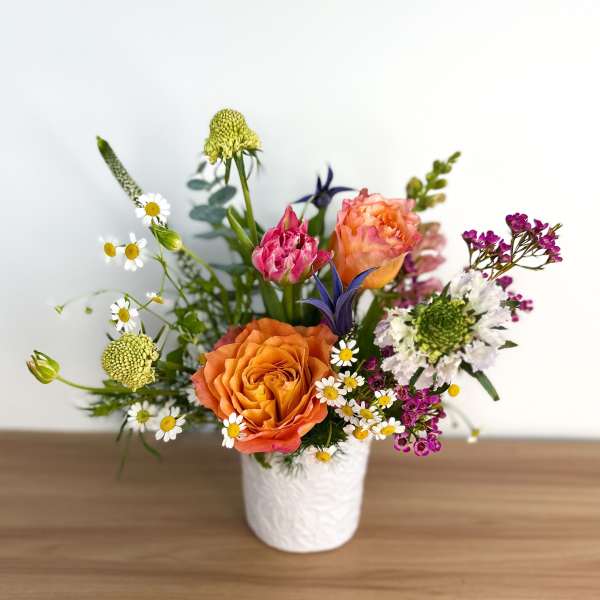 Colorful arrangement of orange roses, pink blooms, and white daisies in a white ceramic vase