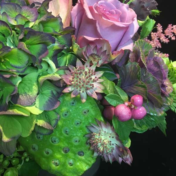 Arrangement of lavender rose, calla lily, hydrangea, berries and lotus pod in rich green and purple tones.