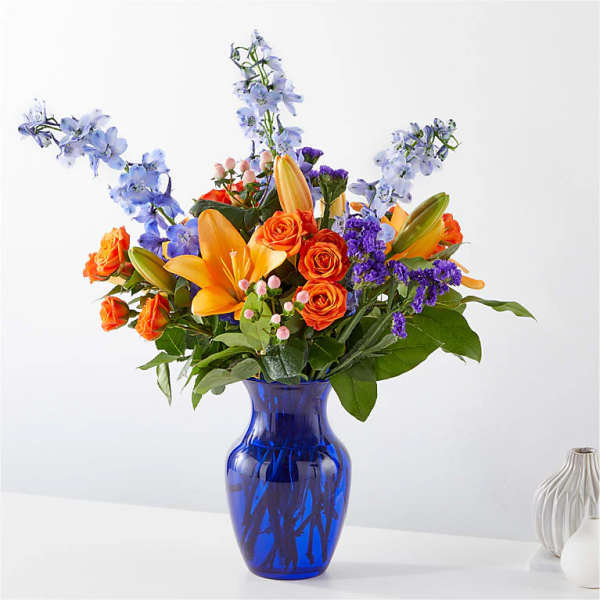 Orange roses and lilies in a blue glass vase with blue flowers
