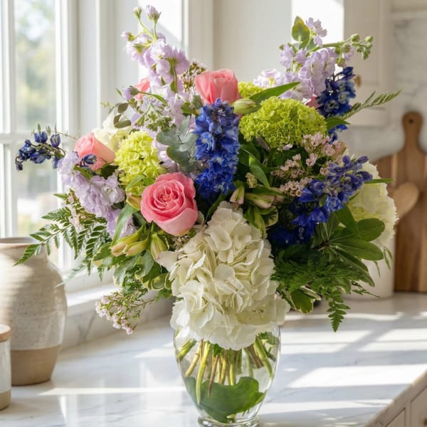 Mixed bouquet of pink roses, white hydrangea, and blue flowers in a glass vase