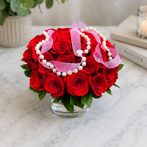 Red roses arranged in a clear glass vase with pink ribbon and pearl strands