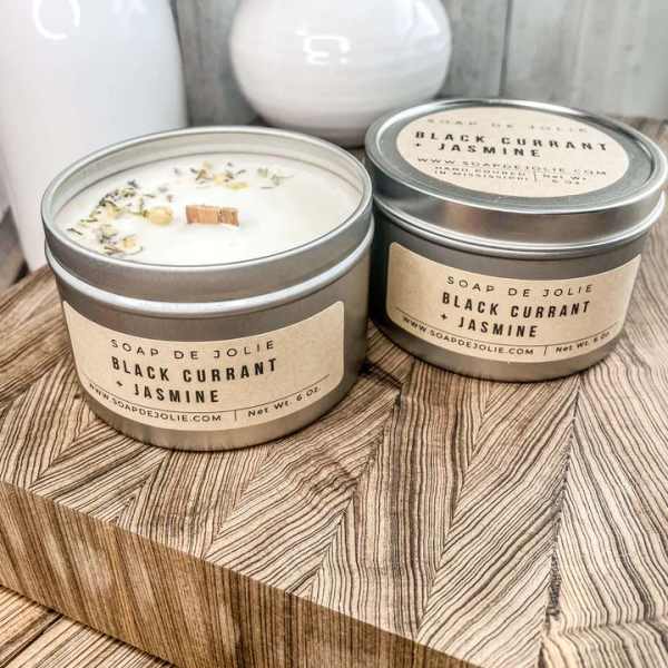 Two lidded tin candles labeled Black Currant + Jasmine on a wood block with white vases behind