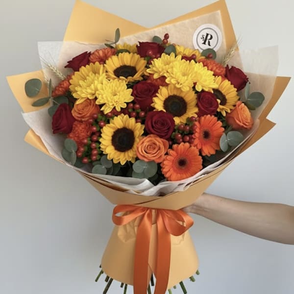 Hand-tied bouquet of yellow sunflowers, red roses, and orange blooms in orange wrap with ribbon