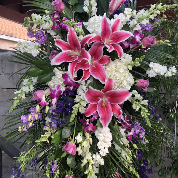 Tall standing spray with pink lilies, white flowers, purple blooms, and palm fronds on a wooden easel