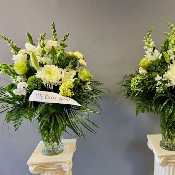Two white and green sympathy flower arrangements in glass vases with ribbon banners
