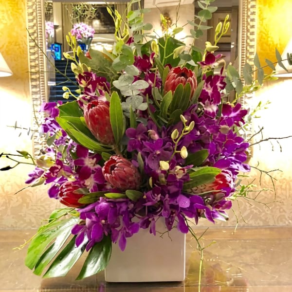 Purple orchids and red protea in a white square vase