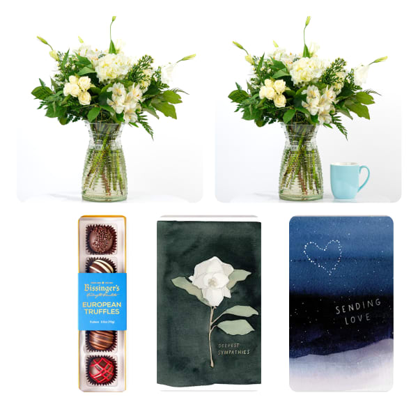 White floral arrangement in a glass vase with a chocolate box and sympathy cards nearby