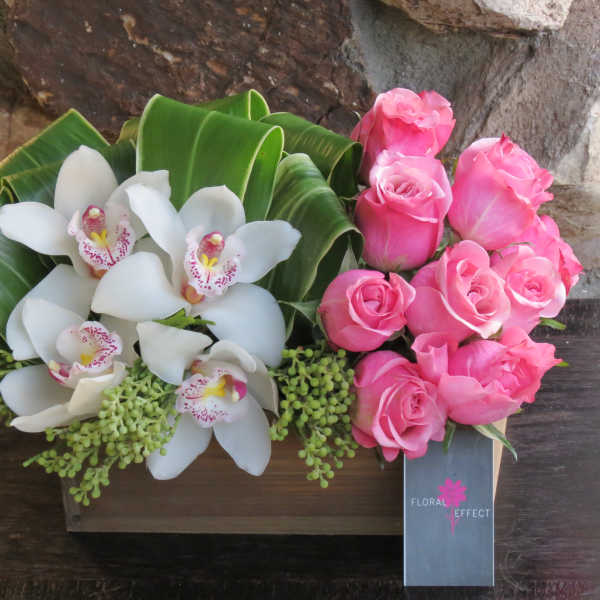 Pink roses and white orchids arranged with broad green leaves in a wooden box