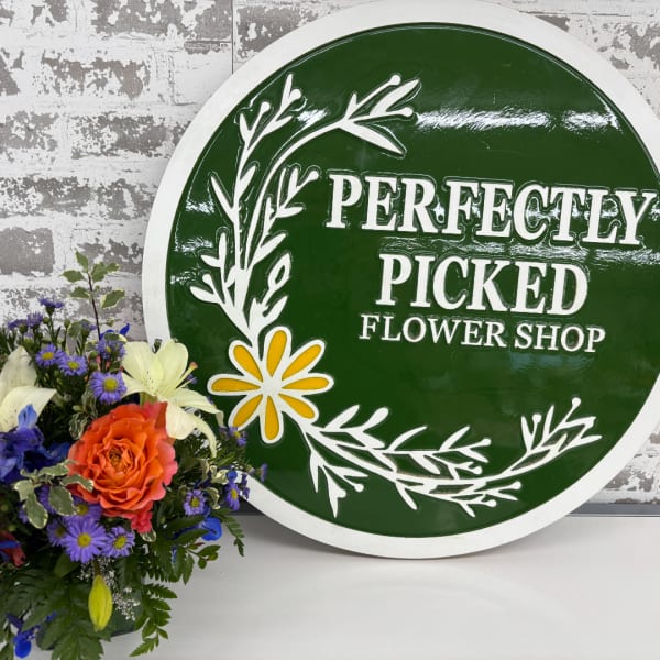Mixed bouquet with orange rose, white lilies, and purple daisies beside a green shop sign