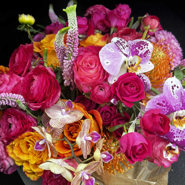 Bright mixed bouquet of pink roses, orchids, and orange blooms in brown wrap