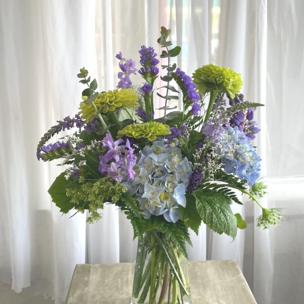 Mixed bouquet of blue hydrangeas, purple flowers, and green blooms in a glass vase
