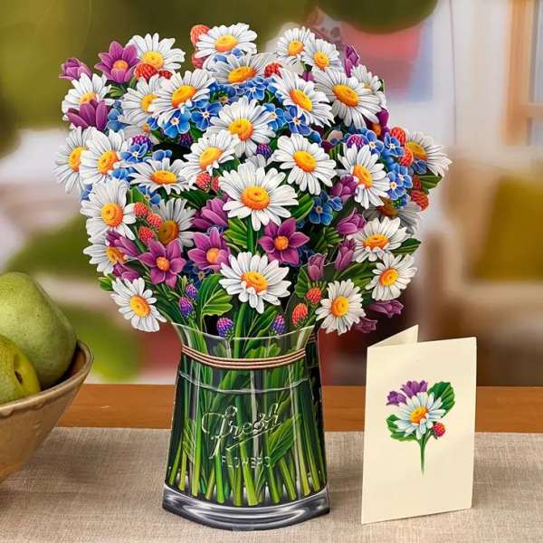 Vase of white daisies with purple and blue accent flowers beside a matching floral greeting card on a table