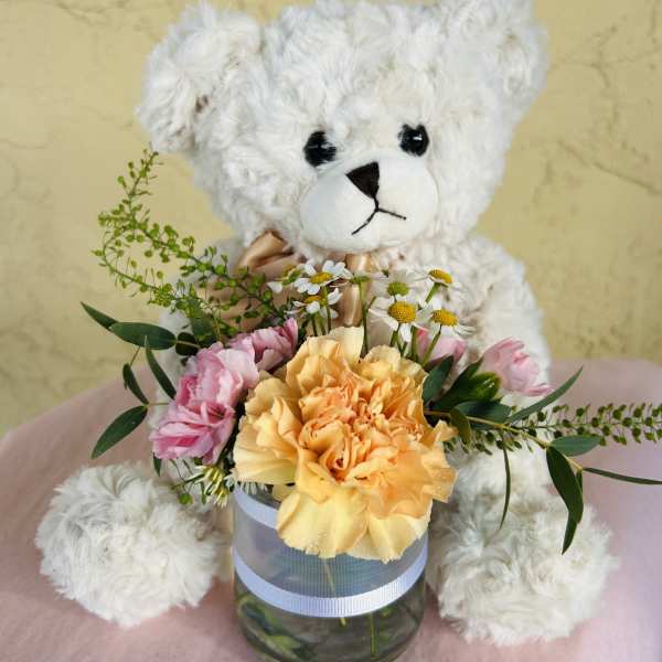 White teddy bear holding a small flower arrangement in a glass vase