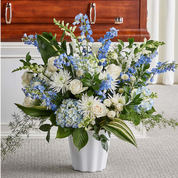 Large blue and white flower arrangement in a white urn in front of a wooden casket