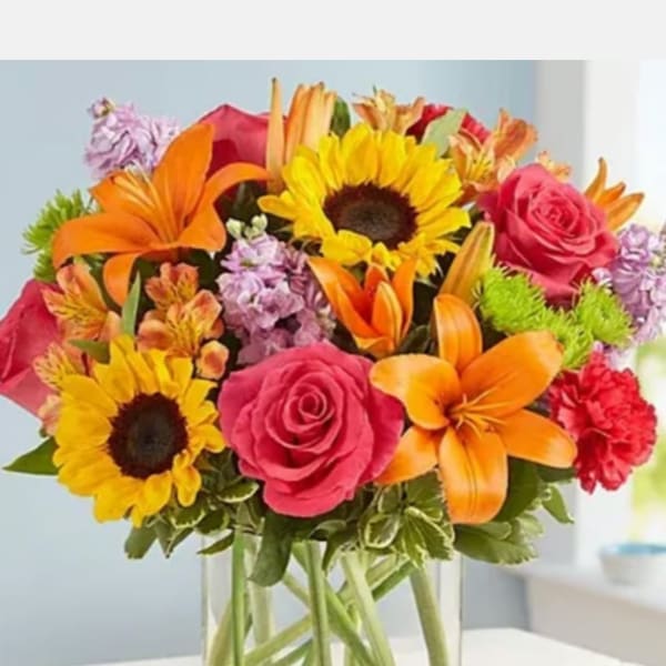 Bright mixed bouquet in a clear glass vase with sunflowers, roses, lilies, and carnations.