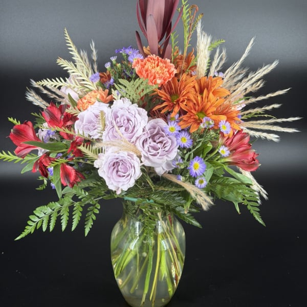 Mixed bouquet of lavender roses, orange mums, and red flowers in a glass vase