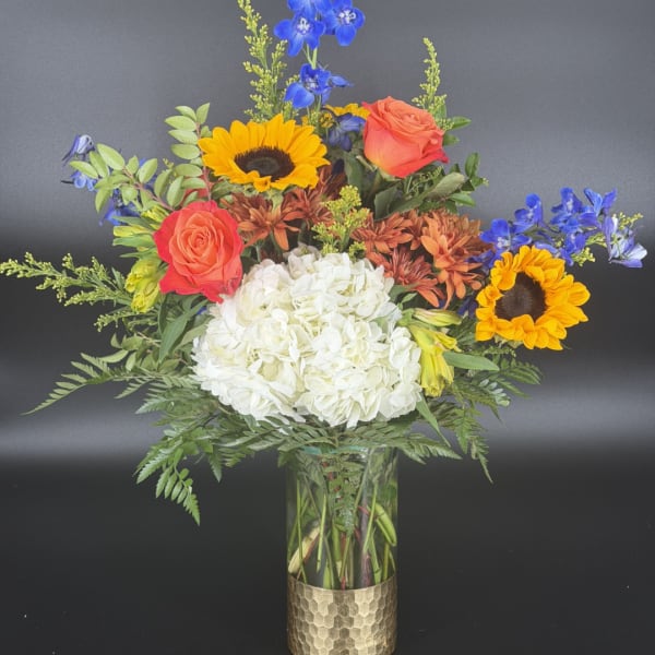 Mixed bouquet with sunflowers, roses, blue delphinium, and white hydrangea in a glass vase