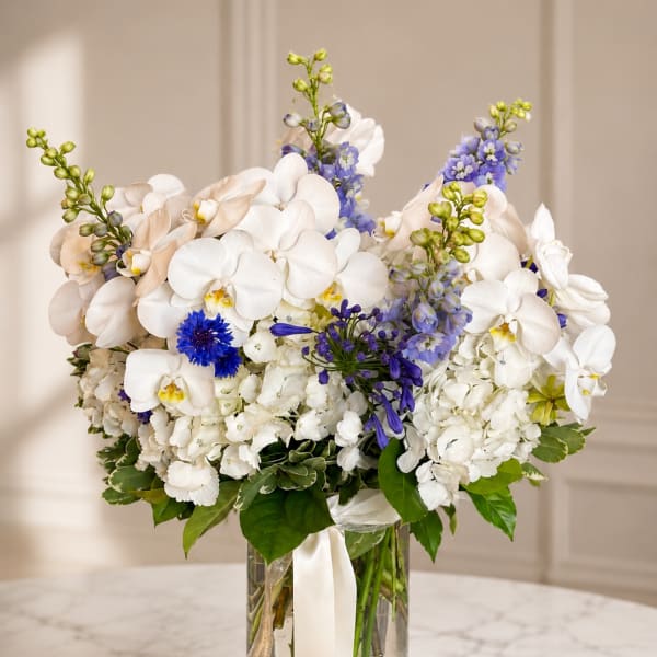 White orchids and blue flowers arranged in a clear glass vase with ribbon