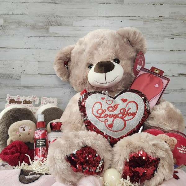Large teddy bear holding a heart pillow with assorted gift items