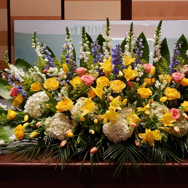 Large stage arrangement of yellow lilies, roses, white hydrangea, and blue delphinium.