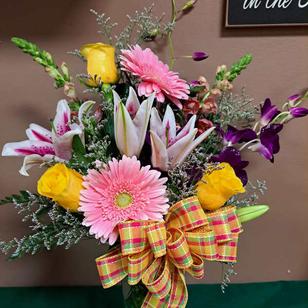 Mixed bouquet of pink gerbera daisies, yellow roses, lilies, and orchids in a glass vase