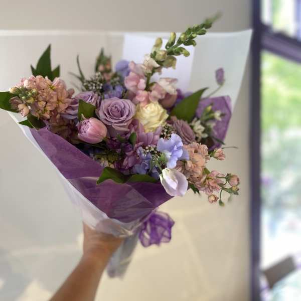 Handheld bouquet of lavender, pink, and cream flowers wrapped in purple paper