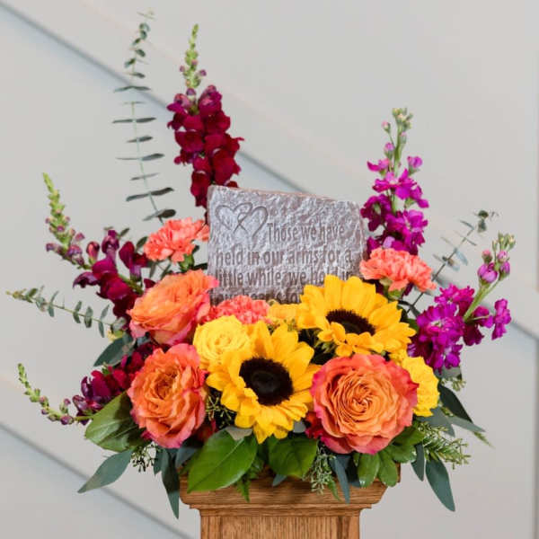Bright mixed flower arrangement with sunflowers, roses, and purple blooms in a wooden container