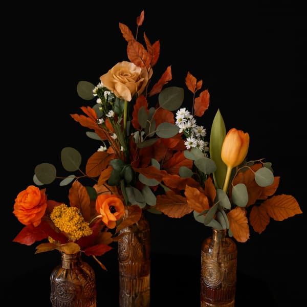 Three autumn floral arrangements in amber glass bottles with orange blooms